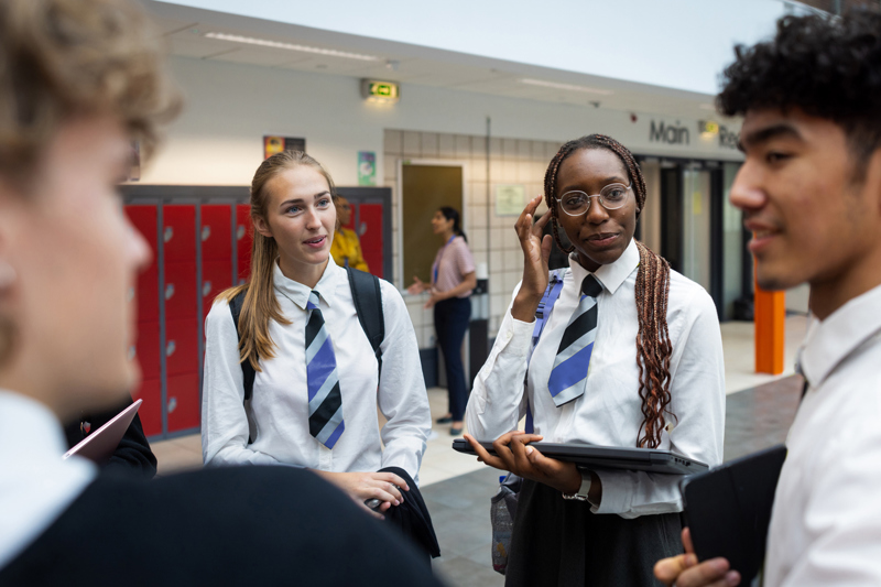 teenage school children holding closed laptops are chatting in a school hallway, wearing school shirts and ties