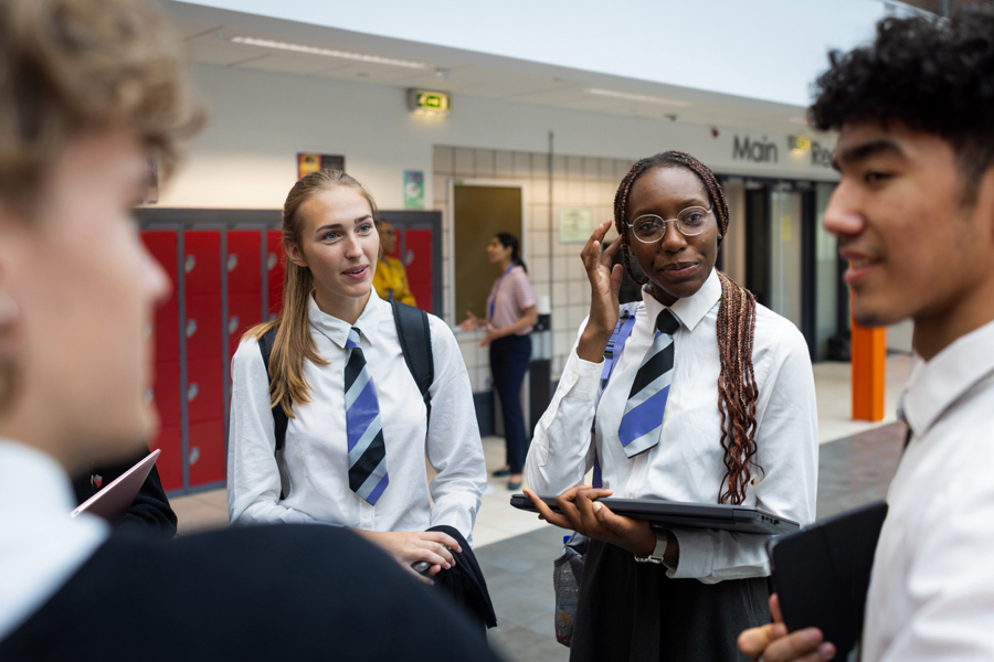 teenage school children holding closed laptops are chatting in a school hallway, wearing school shirts and ties